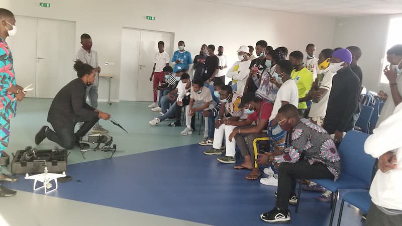 An instructor kneels on the floor with two drones in front of a group of onlooking students